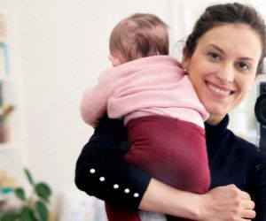 Elena Rossini, holding a camera to take a mirror-selfie with baby on shoulder, bookshelf in background.
